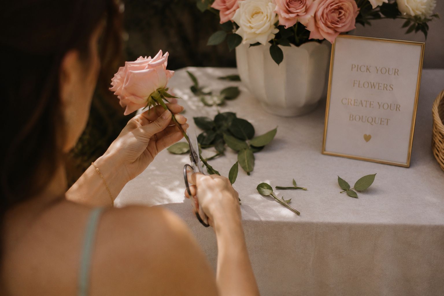 Hands trimming a blush rose over a linen-covered table, beside a framed 'pick your flowers' sign